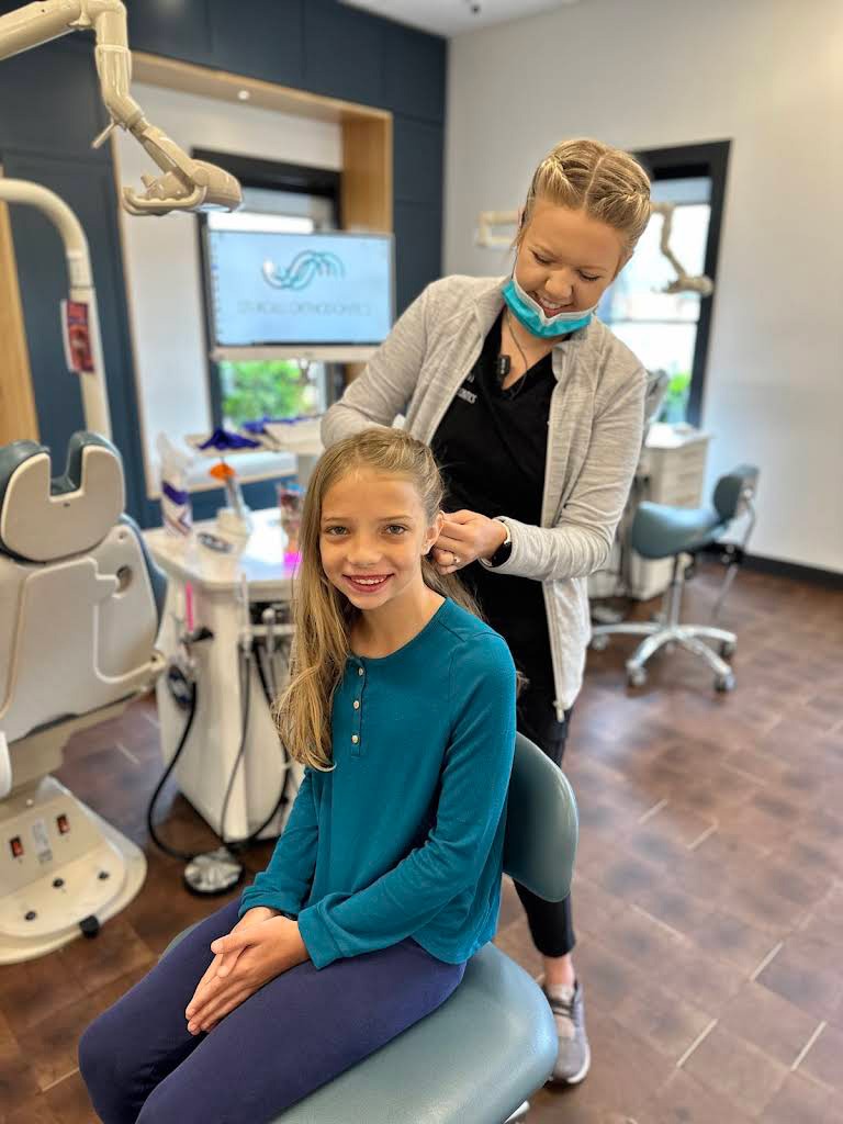 A happy young patient with clear braces smiling in the dental chair at Sturgill Orthodontics while a staff member braids her hair, demonstrating the practice's personalized care and attention to making orthodontic visits comfortable and enjoyable for children.
