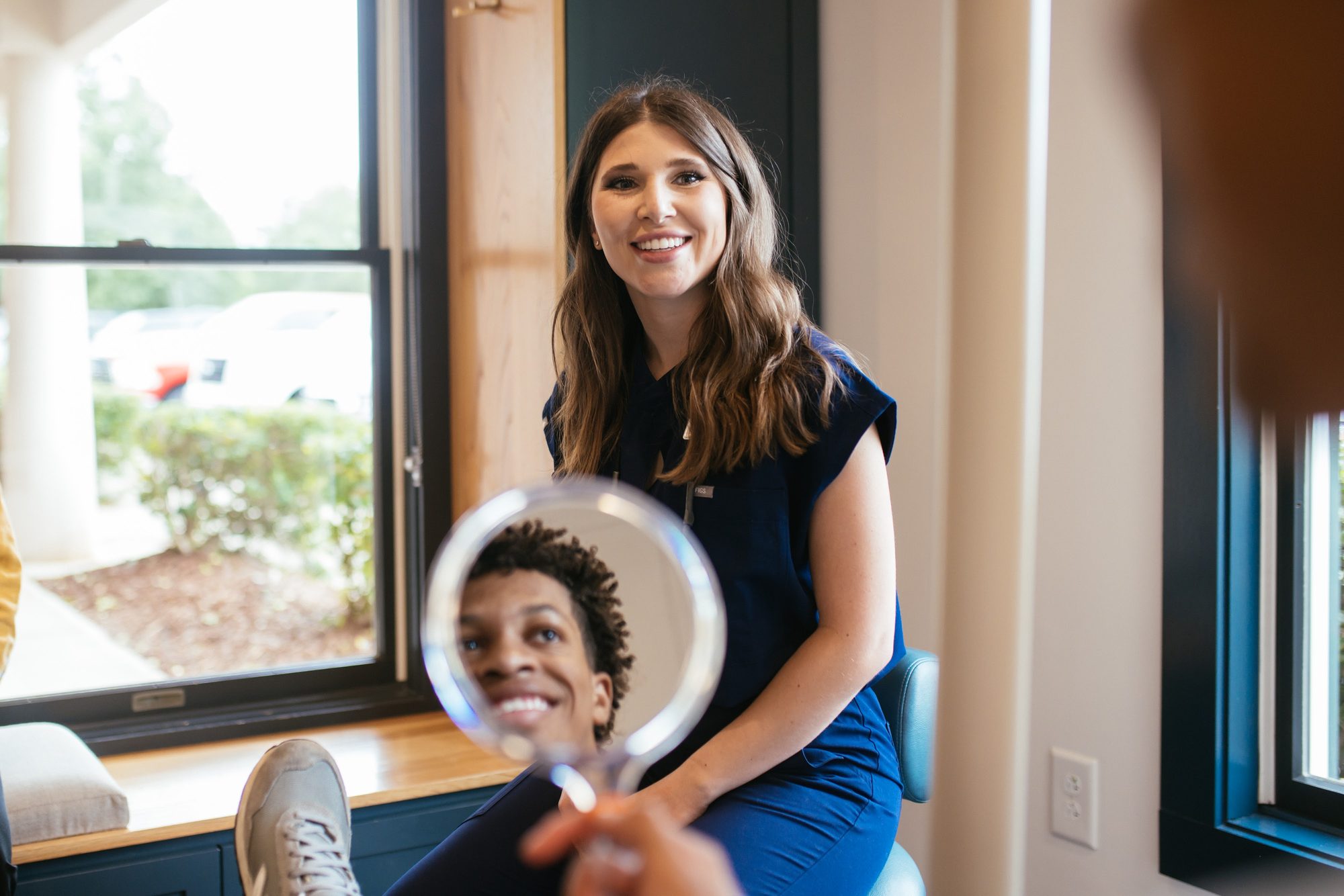 Teen patient smiling into a hand mirror while a Sturgill Orthodontics team member looks on